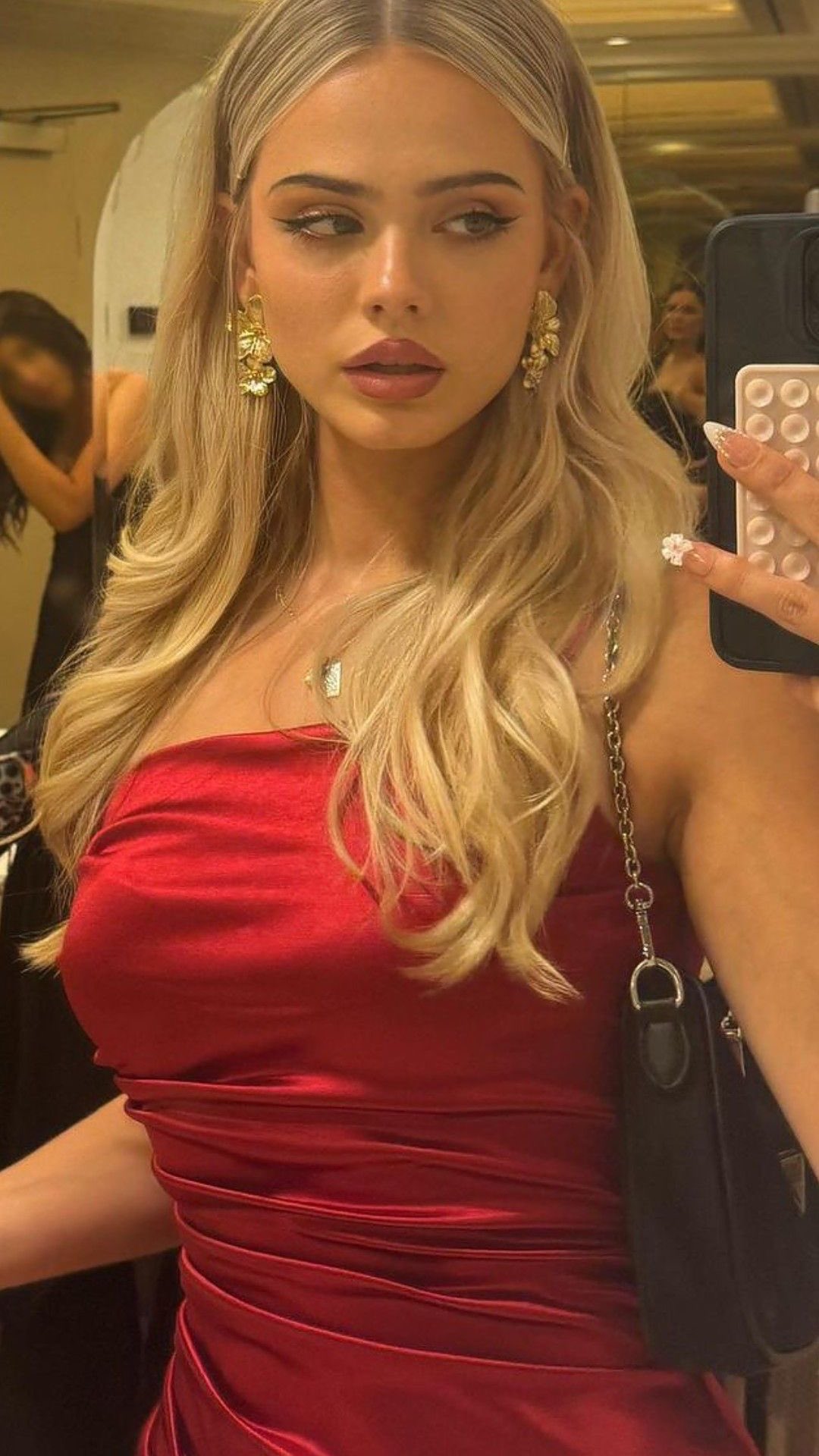 A selfie-style image showing a woman in a red, one-shoulder dress with blonde hair and elegant gold earrings. She is holding a phone to her right with a white floral design on the screen. In the background, other people are slightly blurred, possibly in a dressing room setting. The image uses soft lighting and a neutral background to focus attention on the woman.