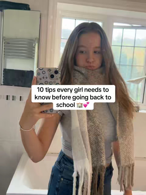 A young woman takes a selfie in a bathroom setting. She is wearing a light gray top, blue jeans, and a patterned scarf. The background includes a mirror, a white bathtub, and a window. Text overlay reads "10 tips every girl needs to know before going back to school".