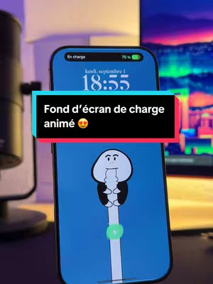 An iPhone displays a custom animated charging screen. The phone is in focus, with a black banner at the bottom containing the title "Fond d’écran de charge animé 🥰". The charging animation features a cartoon figure. The background includes a desk environment with a microphone and monitor.