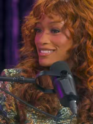 The cover image features a well-lit, close-up shot of a smiling woman with long, wavy auburn hair. She is holding a microphone, suggesting she is likely speaking into it. The background is blurred, but hints at a studio setting. The woman's expression is warm and inviting, and she's wearing an ornate top that adds to the image's visual interest. The composition is well-balanced, with the microphone positioned in the foreground and the woman's face as the main focus.