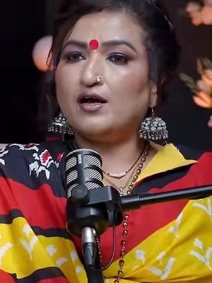A woman is seated in front of a microphone, likely for a podcast or interview. She is fair-skinned, with dark hair styled away from her face, and a red bindi on her forehead. She wears large silver earrings. Her top is composed of black, red, and yellow patterns.