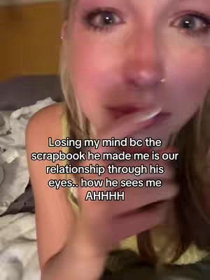 Close-up shot of a young woman, appearing emotional. Her face is centrally framed, with focus on her eyes, which appear teary. She has light skin, blonde hair, and visible eye makeup. The background suggests an interior setting with neutral-toned elements, like fabric/blankets. The lighting is natural, soft, and warm toned.