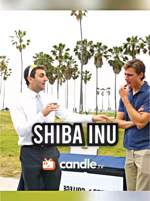 Two men standing outdoors near palm trees are captured in the image. One man, in a white shirt and tie, gesticulates while holding a cup, and a sign that says the title of the video 'SHIBA INU' in front of them. The other man, in a dark blue polo, appears to be contemplating or shocked. The image contains a website title, and the overall setting suggests an outdoor event or presentation, with a backdrop of tall palm trees.