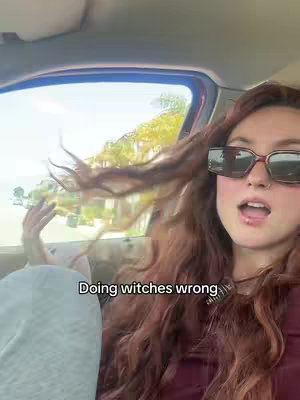 A young woman with long auburn hair wearing sunglasses is looking slightly to the right. The car window is visible behind her, with sunlight and foliage in the background.