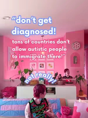 A selfie-style shot in a bright pink room. The person in the foreground, with short pink hair and tattoos on their arm, is looking towards the left with a neutral expression, and there's a pink stuffed animal on a couch in front of them. The background has framed pictures with a floral pattern. Text overlays are present.
