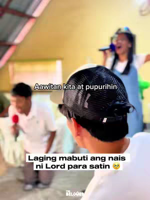 A person wearing a cap is in the foreground, facing away from the camera. In the background, two people are visible, one holding a microphone and singing. Text overlays are present, with the main text in white against a partially blurred background.