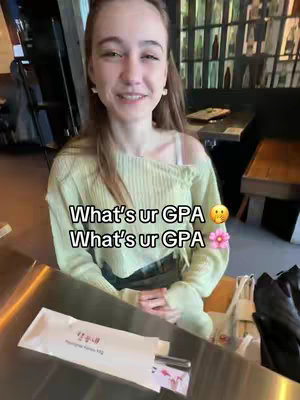 A woman is taking a selfie while seated in a restaurant. Overlay text asks "Whats ur GPA" in two rows. She wears a light green sweater with a white undershirt. A white pack of utensils and a napkin are in the background.