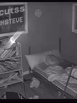 The image is a black-and-white, low-quality surveillance-style shot of a messy bedroom. The room is dimly lit, with the main focus on a bed covered in rumpled sheets and pillows. A bookshelf on the left holds a collection of books and papers, partially illuminated by a lamp. On the window, there is text. The overall composition gives a sense of disarray and a voyeuristic perspective.