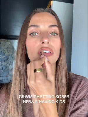 A close-up shot of a woman applying lip gloss. She is looking directly at the camera, and the focus is on her face and hand. The background is blurred, but a mirror frame and part of a wall are visible.