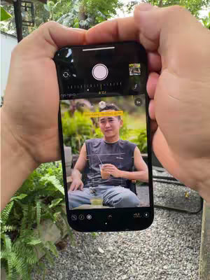 A person's hands hold an iPhone showing a portrait being taken. The screen of the iPhone displays a man holding a cup and sitting outside. The background is blurred green foliage and a pebbled path, suggesting a café or outdoor setting.