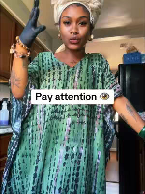A woman in a green patterned dress is looking at the camera with a serious expression. She is wearing a white head wrap and various accessories. Overlaid text reads "Pay attention" with an eye symbol.