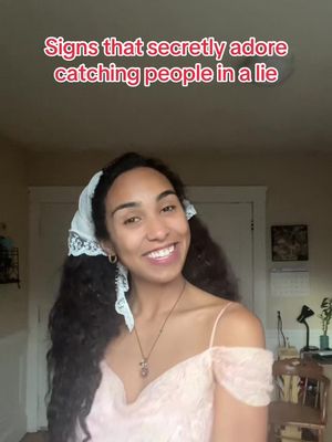 A woman with dark, curly hair smiles at the camera. She wears a light pink dress with off-the-shoulder straps. There's a white scarf on her head and a necklace. Text at the top says, 'Signs that secretly adore catching people in a lie'.