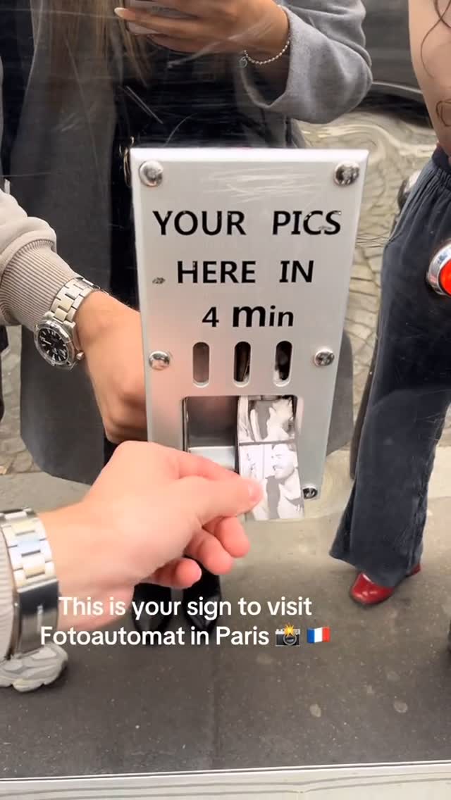 A person's hand reaching into a vintage photo booth to retrieve a strip of black and white photos. The machine is silver with text that says 'YOUR PICS HERE IN 4 MIN.' Two people are visible in the background, one wearing a grey overcoat, and the other wearing black pants and red shoes. The floor is dark grey.