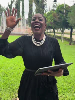 A woman stands outdoors with a park-like background, smiling widely and waving with one hand. She wears a black dress, a pearl necklace, and a bracelet. She is also holding a tablet. The background features a fountain and trees, and the lighting appears to be natural.