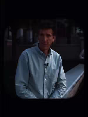 The cover image presents a man in a light blue button-down shirt, speaking. He is positioned in front of a partially visible, outdoor background. The composition is framed within a darker, somewhat rectangular border, and includes a microphone clipped to the man's shirt.