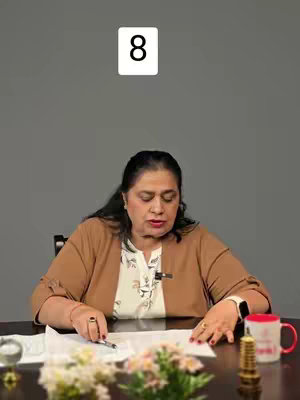 A woman sits at a table, facing the camera. White text box with number 8 is the top center.  She is looking down at papers in front of her. Behind her is a grey background.