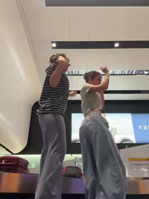Two young women dance spontaneously in an airport setting, with their arms raised in the air. The woman on the left is wearing a striped shirt and grey pants, and the woman on the right is wearing a crop top and pants. The background includes airport fixtures and a large screen.