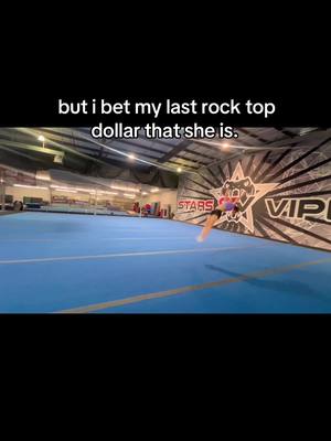 A young person in a cheerleading uniform is mid-air, performing a tumbling skill in a cheerleading gym. The background includes blue mats, gym equipment, and a wall with the team's logo and a sponsor's branding.