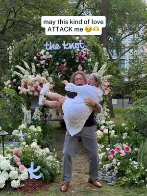 A man holding a woman up in his arms in front of a flower-adorned structure with the words 'the knot' in neon lights, surrounded by lush greenery and flowers; the couple's facial expressions convey joy and excitement, indicating a wedding or celebration.
