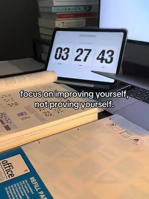 The image shows a desk setup with a digital timer on a tablet displaying the time "03:27:43".  Below, open books and paper refills create more workspace on the desk. In the background, more books are stacked, a computer monitor, and a pen.