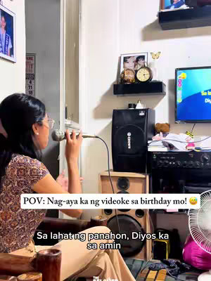 A woman wearing glasses and a patterned shirt sings into a microphone in a room. Behind her are speakers, a TV screen displaying karaoke lyrics, and decorative items on a shelf. The layout suggests a home karaoke setup, reflecting a lively atmosphere.