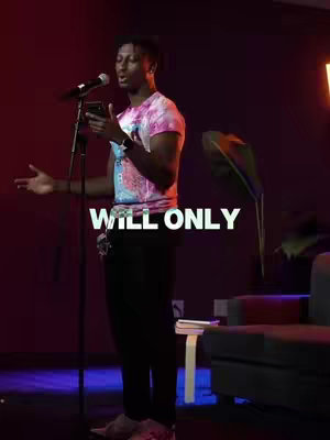 A man in a tie-dye shirt stands angled to the camera, holding a phone and microphone. The background is a blurred mix of dark red and subtle lighting.