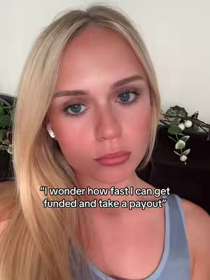 A young woman with blonde hair and light makeup looks at the camera with a slight frown. The background is out of focus and includes dark furniture with greenery.