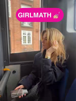 A woman with blonde hair sits on a bus, looking out the window. A pink banner with the text "GIRLMATH" is overlaid at the top. The background includes a brick building and a window. She has a phone in her hand and is wearing a black jacket.