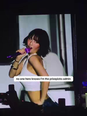 A woman with short black hair, wearing a white crop top and dark bottoms, is singing into a purple microphone. The backdrop features an indistinct bright light. A text overlay reads, 'no one here knows I'm the prizepicks admin'.