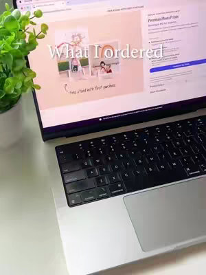A laptop displaying a website, with a bright green plant to the left and a black keyboard at the bottom. The laptop screen features the words "What I ordered" over an image and text related to photo prints.