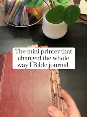 The cover image shows a close-up of a maroon Bible open on a desk. A small white sticker displaying the hook text is overlaid on the upper part of the Bible. In the background is a clear container of colored markers, a small succulent in a white pot, and a black desk surface.