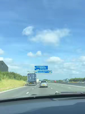 A first-person perspective view from inside a car, looking forward on a highway. A blue, cloudy sky dominates the top portion of the image, with a highway sign visible in the middle distance, showing place names. Trees run along the side of the road.
