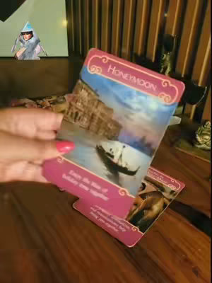 The cover image is a medium shot of a person holding a tarot card that says "Honeymoon". The card shows a canal with a boat and some buildings. There is another card below it. The person is holding the cards in front of a wooden table background. There is a small image in the top-left corner of a person's face. There are other vertical decorations in the background.