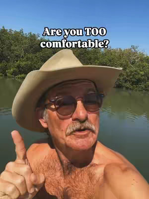 A man with a hat, sunglasses, and no shirt is pointing with his right hand in front of a river with trees and blue sky in the background, with text overlayed at the top that says "Are you TOO comfortable?" in white text.