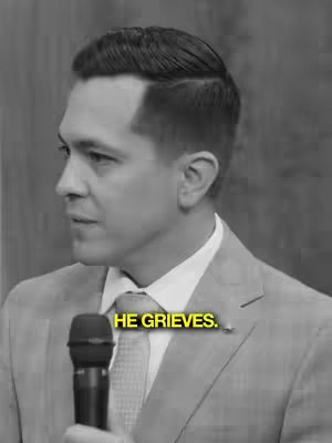A black and white image featuring a man wearing a suit, seen in profile from the shoulders up. The man is addressing someone off-camera, holding a microphone. Text overlayed on image: HE GRIEVE.