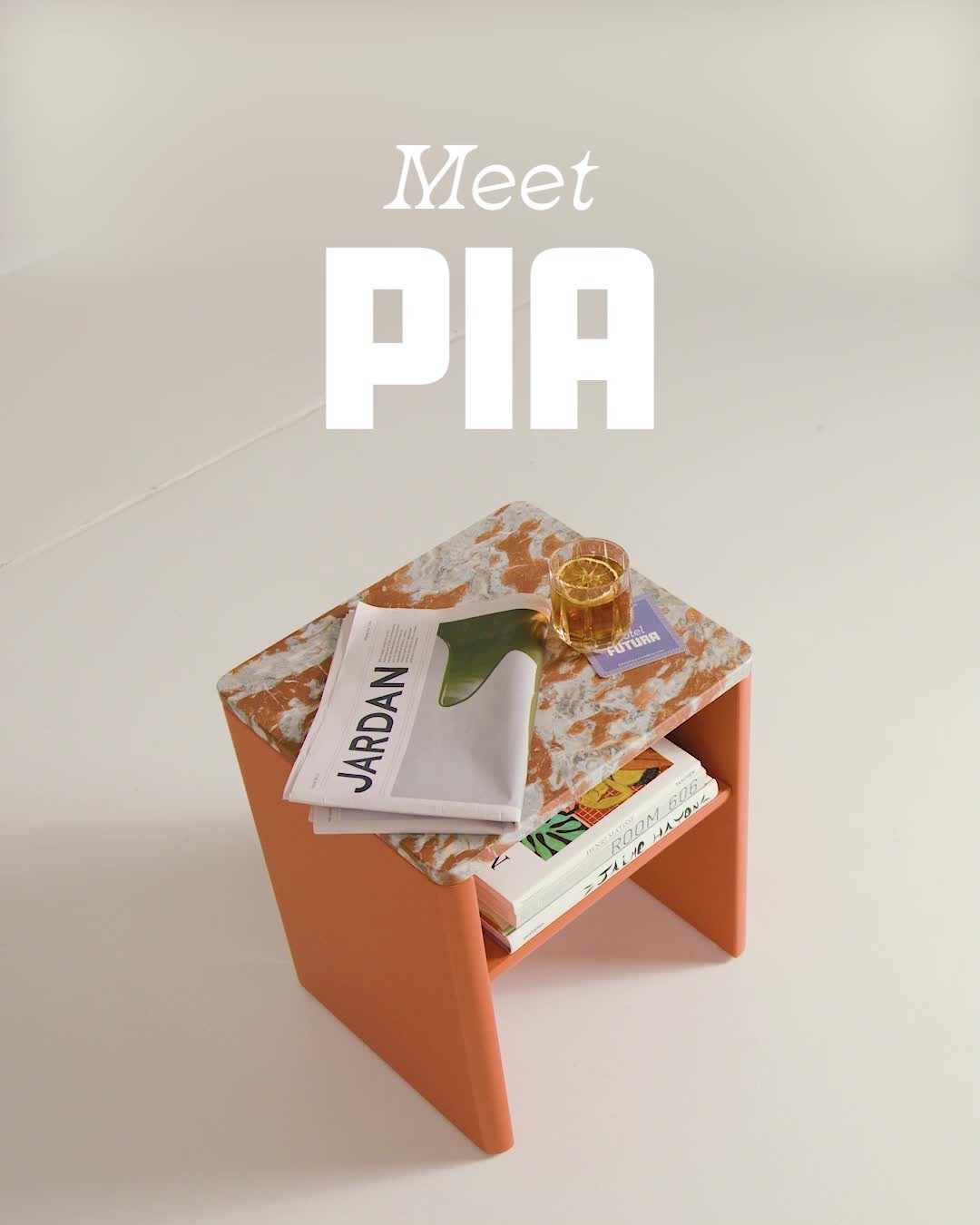 A high-angle shot presents a modern side table, 'Pia', against a neutral beige backdrop. The table's structure has a bright orange base, supporting a top with a marbled, speckled design in white, brown and tan. The table features an open shelf, holding books and a magazine while a glass with an orange in is also on top.