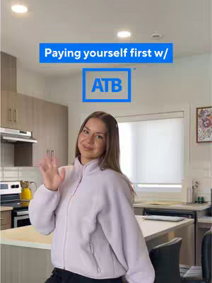 A young woman is standing in a kitchen, smiling and waving. Above her, centered, is a rectangular blue shape containing white text that reads, "Paying yourself first w/." Centered below that, also enclosed in a blue rectangle and outlined in white is the letters "ATB." The lighting in the kitchen is bright and appears to be from overhead. The background includes light-colored cabinets, a window, and appliances.