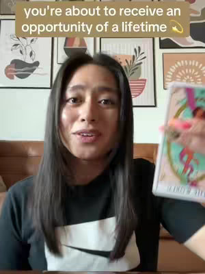 A person with long dark hair is holding a tarot card, smiling into the camera. A text overlay at the top says 'you're about to receive an opportunity of a lifetime.' Background includes framed artwork and a brown couch.