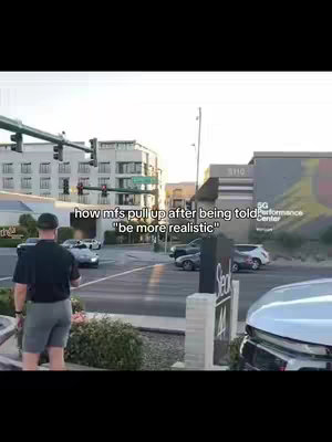 A wide shot of a city street corner features a man in shorts and a dark shirt watching a car stopped at a red light. Across the street, there are several buildings.