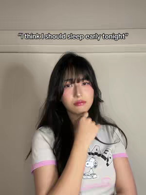 A woman with long black hair and bangs gazes at the camera as she touches her face with her hand. She is wearing a t-shirt with a Snoopy graphic and pink trim. The background is a white wall.