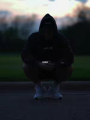 A person wearing a dark hoodie is squatting low to the ground in front of a blurry, outdoor setting at dusk. The figure is silhouetted, obscuring facial features, and the hoodie has a hood up. Text reading "TIMES" is overlaid near the center of the frame.
