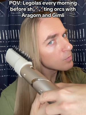 A close-up shot of a person styling their long, blonde hair with a hair styling brush, appearing to mimic Legolas from Lord of the Rings. The person is fair-skinned, with light eyes, and is wearing a green shirt against a patterned, dark blue background. Text overlayed on the image reads, 'POV: Legolas every morning before shooting orcs with Aragorn and Gimli.'