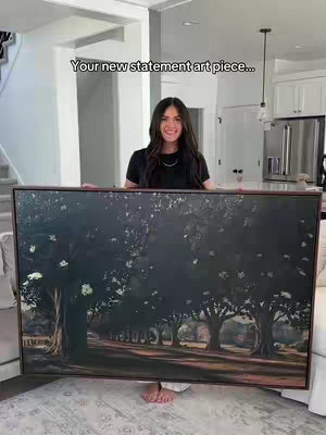 A woman is standing in front of a large framed canvas painting. She is holding the painting in front of her torso, covering the bottom half of her body. The painting depicts a dark road lined with trees under a cloudy sky. The woman has long dark hair and is wearing a black shirt and white pants. The background is a modern home interior.