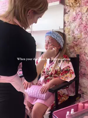 The cover image shows a young girl sitting in a makeup chair, wearing a floral robe and a blue headband. An adult is applying makeup to her face. The background features a wall decorated with pink and white flowers. The overall tone is light and feminine.