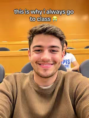 A young man with brown hair and a brown sweater is smiling, taking a selfie in a classroom setting. The background shows wooden classroom seats. On the top of the frame, the overlay text reads, 'this is why i always go to class with a crying emoji.'