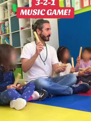 A scene inside a classroom. A fair-skinned man with a beard and his hair tied up in a bun interacts with a group of preschoolers. They're seated on a colorful rug with musical instruments, suggesting an active music class. An overlay in the background states the game's name.