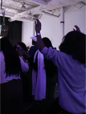 The image shows a group of people with their arms raised in a room. The lighting is dim and purple, creating a sense of reverence or possibly a performance setting. Some individuals are wearing white clothing. The composition focuses on the people from the waist up, suggesting a visual emphasis on their emotional display.