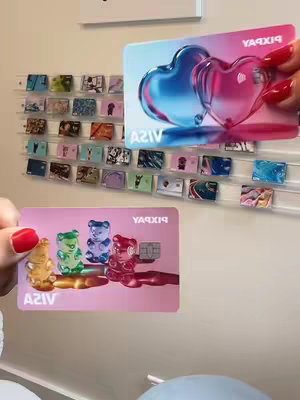 The image shows two Pixpay credit cards being held up. One card, held with a hand with red nail polish, displays two interconnected hearts with a blue to pink gradient, above the word "VISA" below. The other card, below, is light pink and shows four gummy bear shapes in different colors above the word "VISA". Behind the cards, a display of various cards is mounted on the wall.