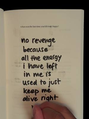 An open book is shown with handwritten text occupying a large portion of the frame. The book is open to a page with a somewhat aged, off-white tone. The text, written in a thick black marker, is the primary focus, arranged in a staggered vertical layout that dominates the center of the image. A hand, with skin tone visible, is partially visible at the bottom.