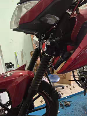 The image shows a close-up of a red motorcycle being worked on. The front end is visible with the headlight, front forks, and part of the wheel. The background is a workshop, with tools and other items suggesting repair work.